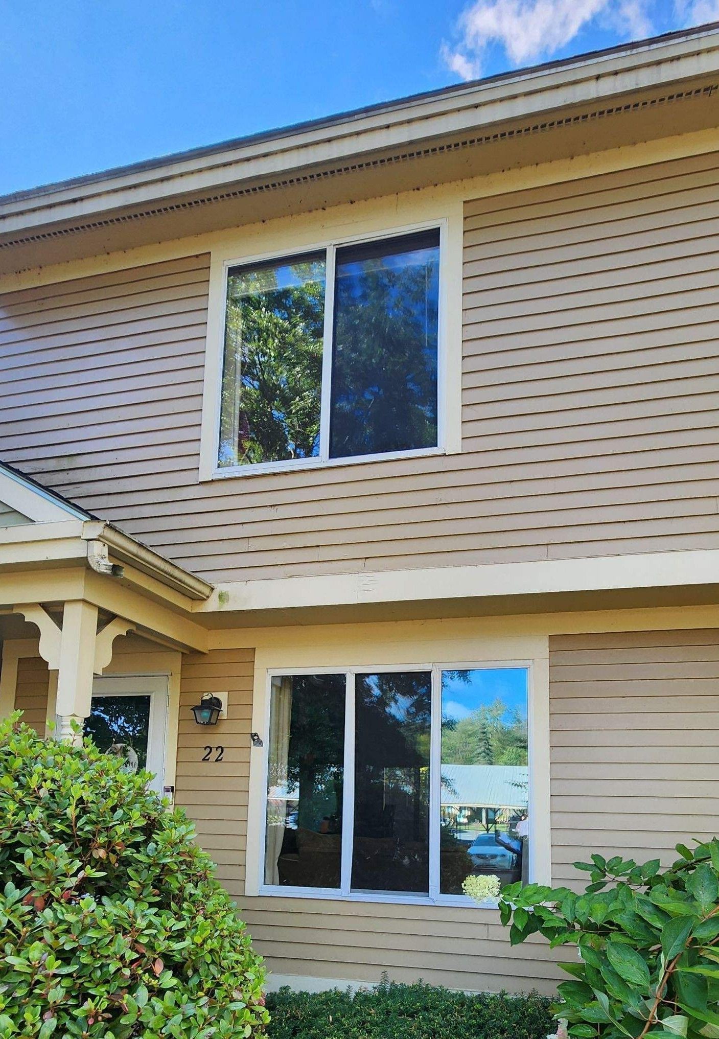 Two-story tan townhouse with white-trimmed windows, front shrubs, and a partial view of a balcony and walkway