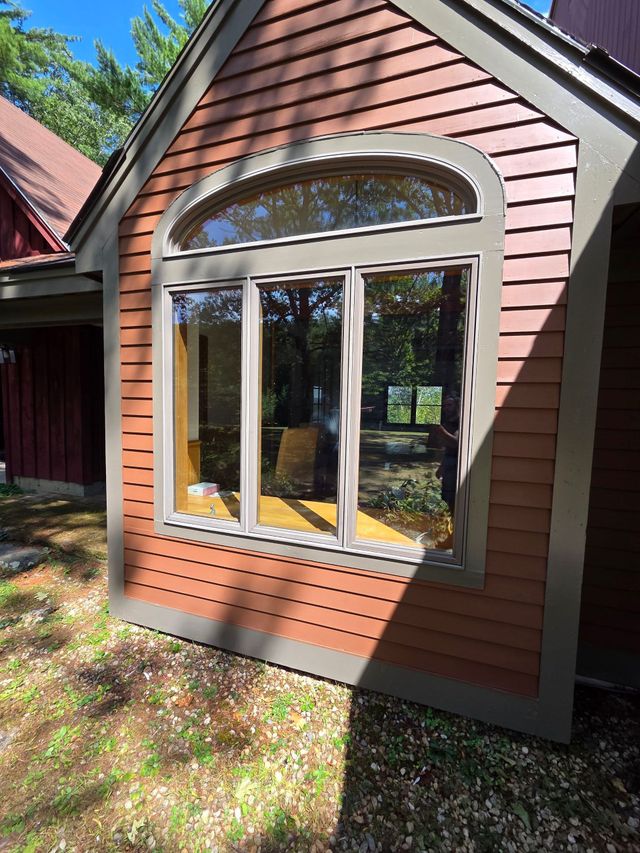 Brown-sided house exterior with a large arched window and white trim in sunlight
