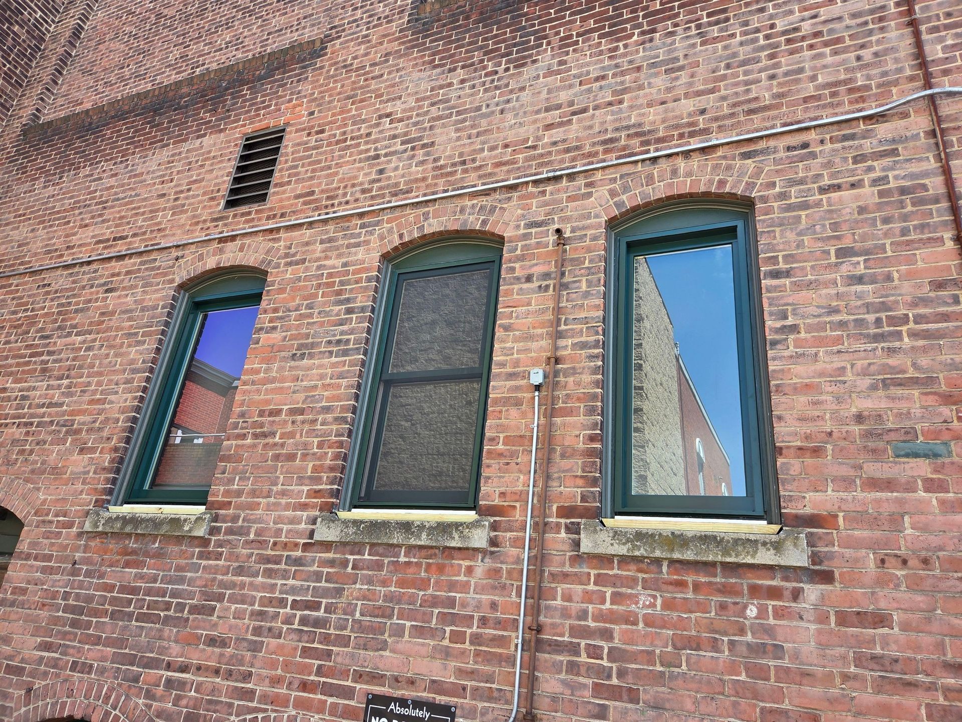 Brick building wall with three green-trimmed windows, one with a blind and two reflecting sky.