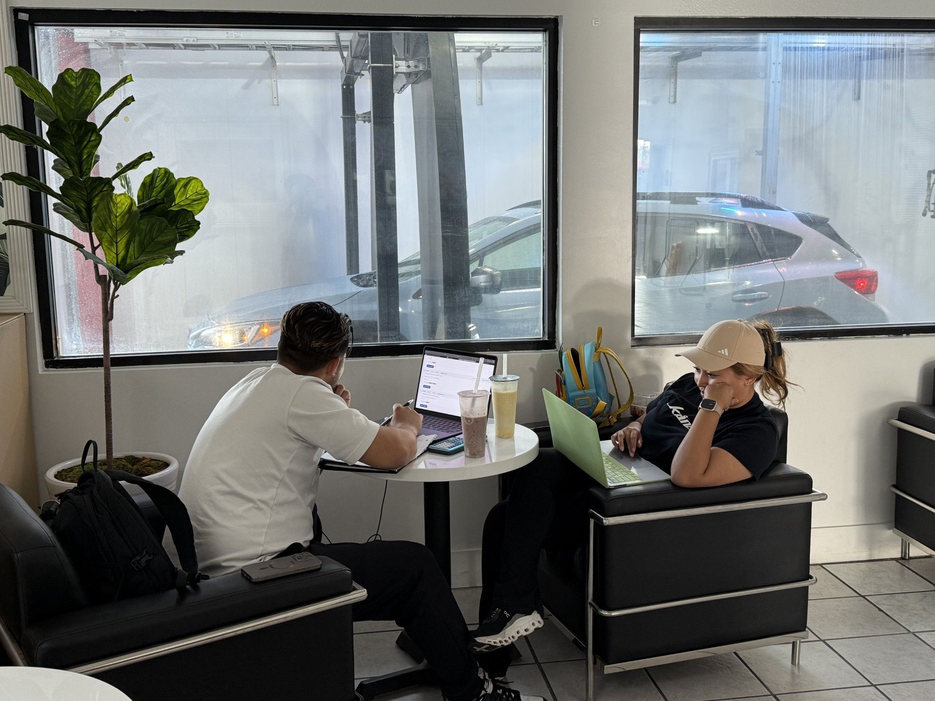 Two people work on laptops at a table in a cafe with a view of a car wash.
