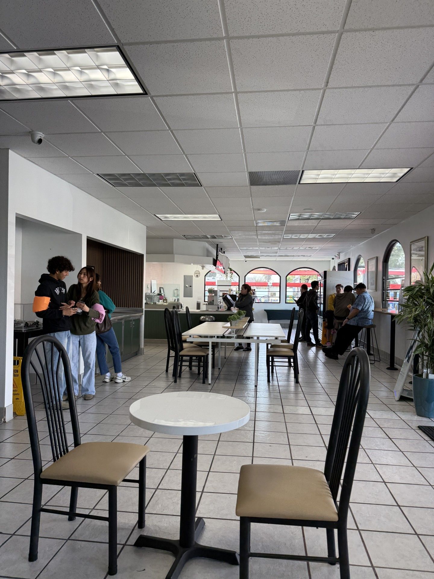 Interior of a restaurant with tables and chairs. People are gathered, and some are seated. Bright lighting and tiled floor.