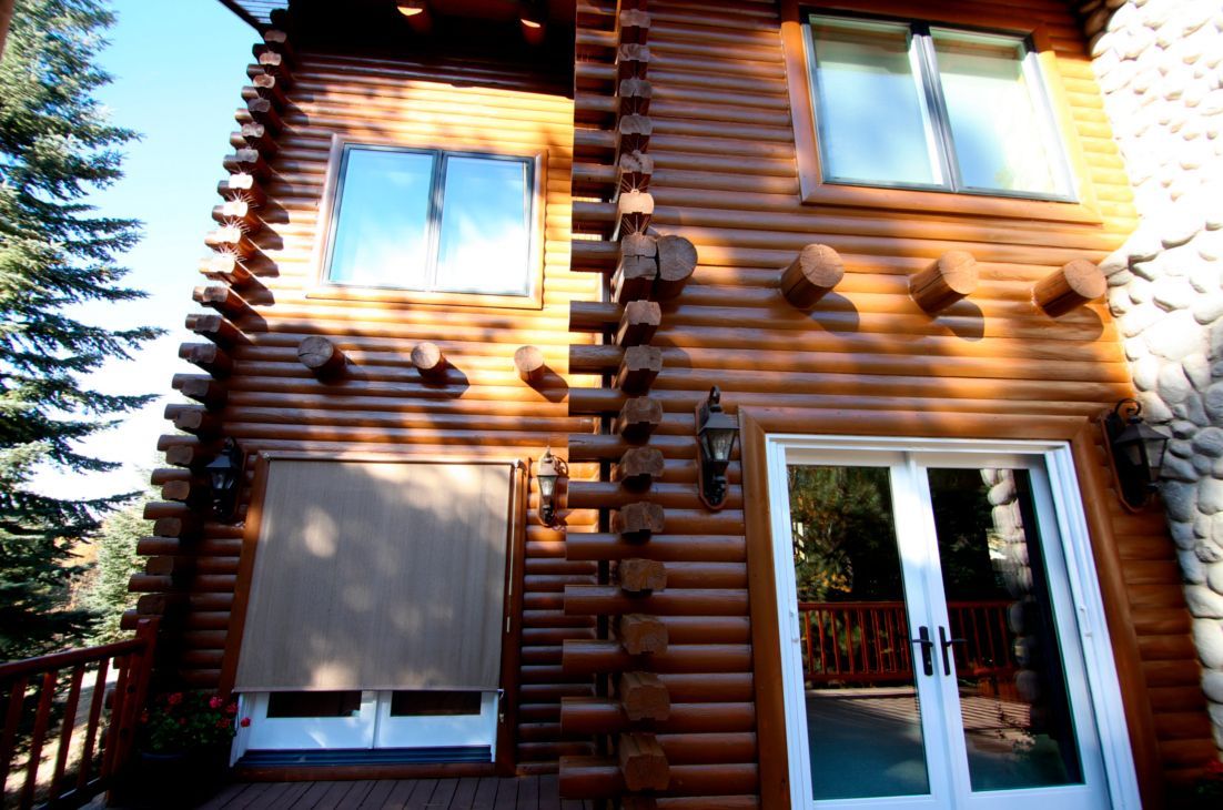 Two-story log cabin with windows, a deck, and French doors. Brown logs and a blue sky.