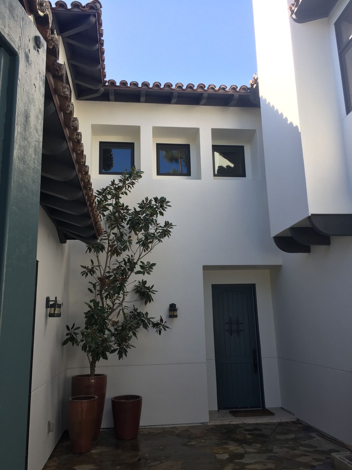 Courtyard with white walls, arched doorway, three small windows, terracotta pots, tree, and blue door.