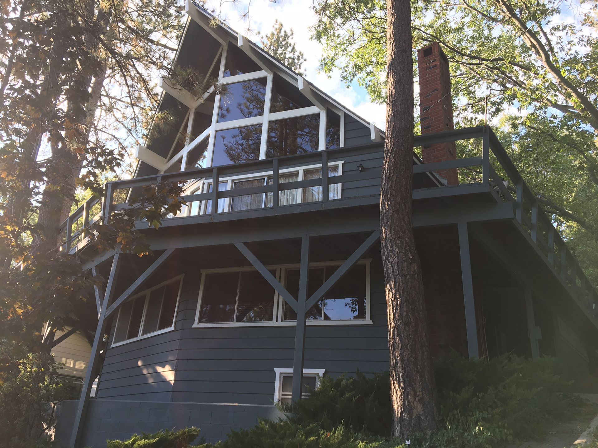 Two-story A-frame house, gray exterior, large windows, deck, chimney, surrounded by trees.