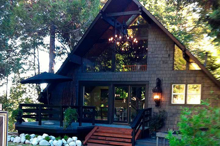 A-frame cabin with dark siding, large windows, and a porch, nestled in trees.