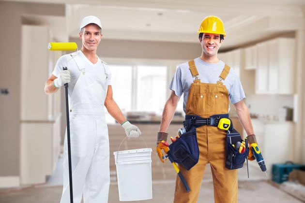 Two construction workers in uniforms and hard hats stand smiling in an interior room, one holding a paint roller and bucket.