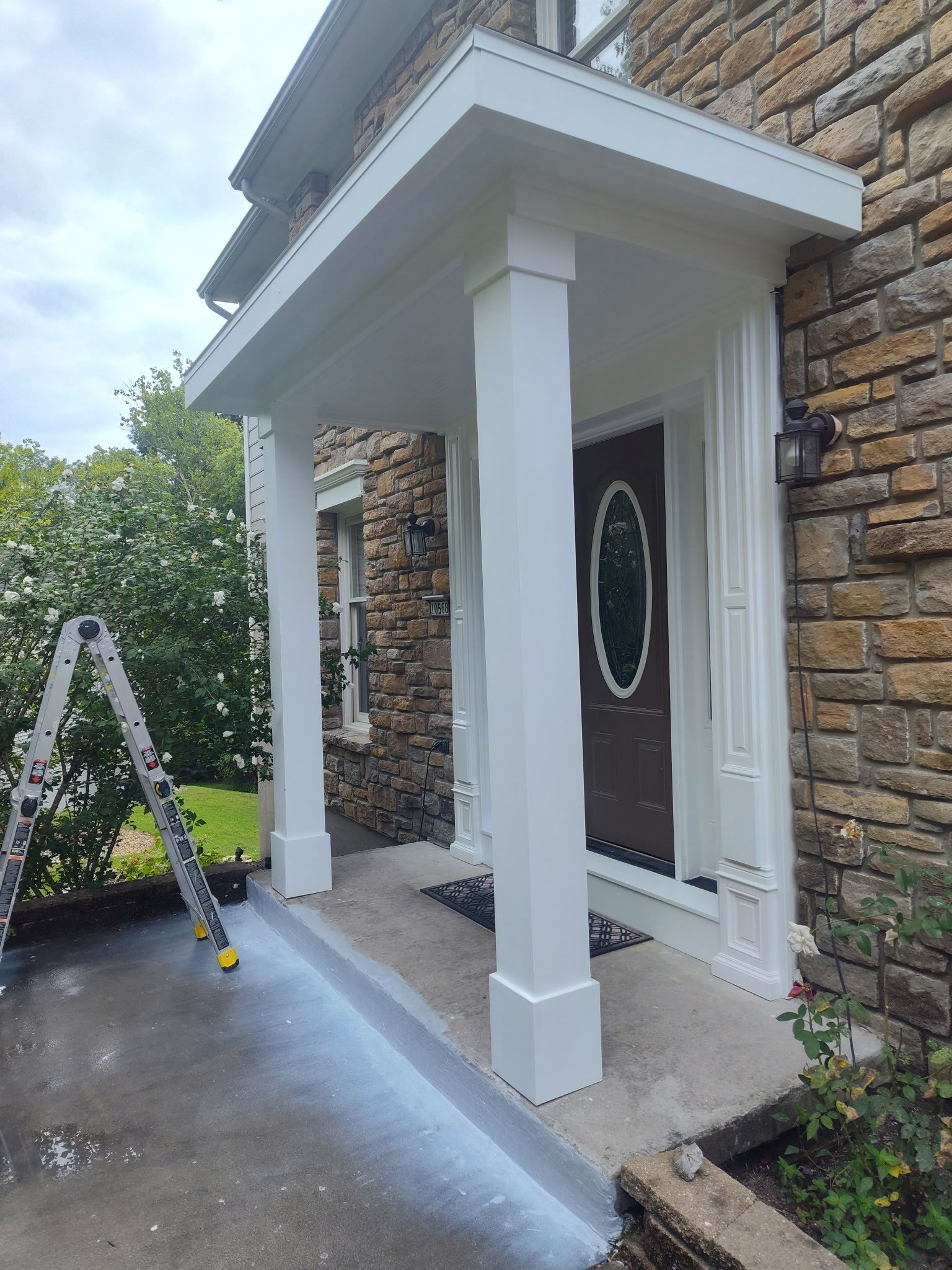 Front porch with white columns, black front door, stone wall, and a step ladder beside the entryway.