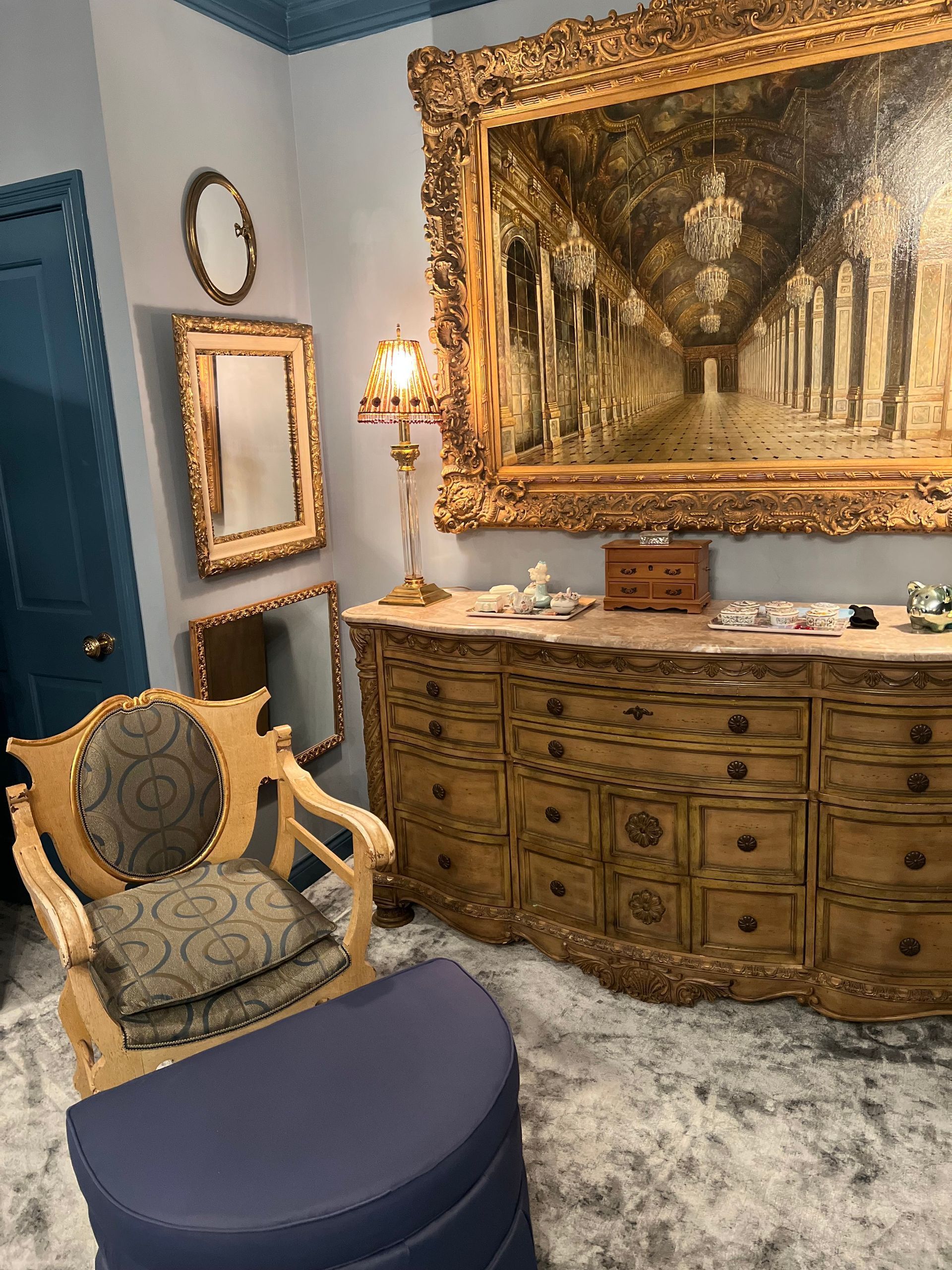 Elegant living room with ornate gold-framed painting, vintage chair, and curved wooden dresser on a plush rug