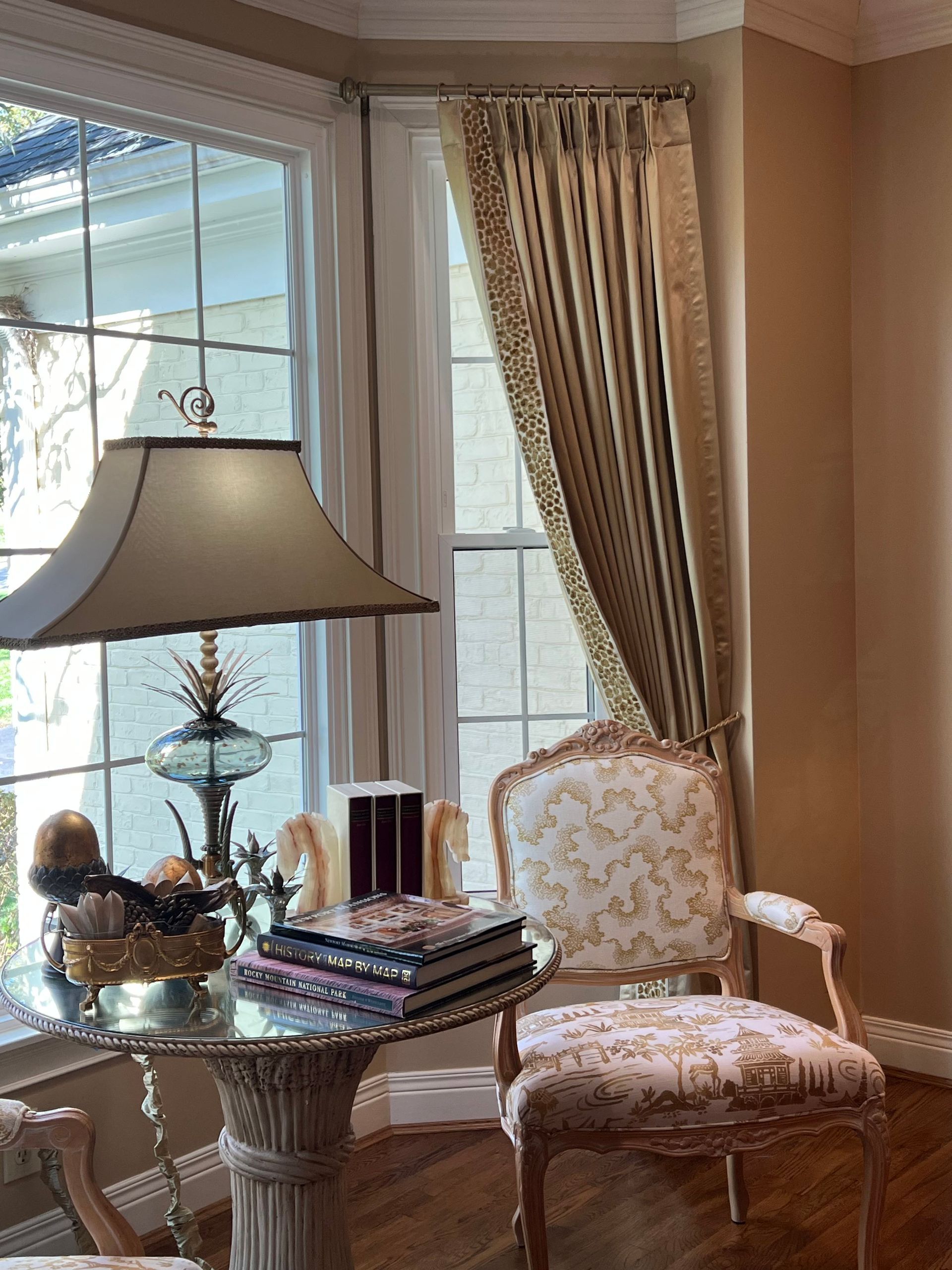 Elegant sunlit sitting room with floral chair, lamp, draped curtains, and a table with stacked books and decor.