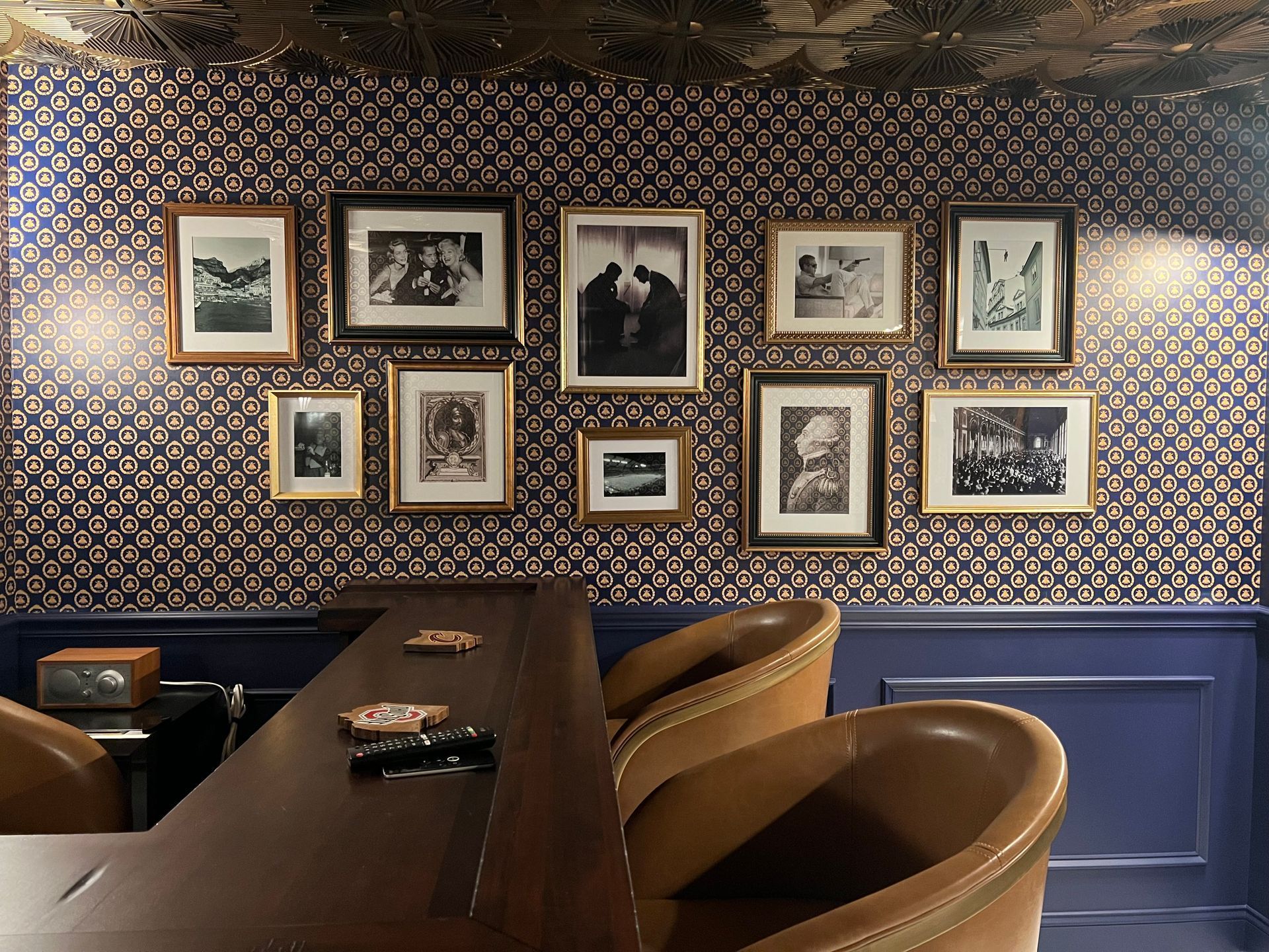 Cozy restaurant booth with a dark wood table, tan chairs, blue wainscoting, and a wall of framed art and patterned wallpaper