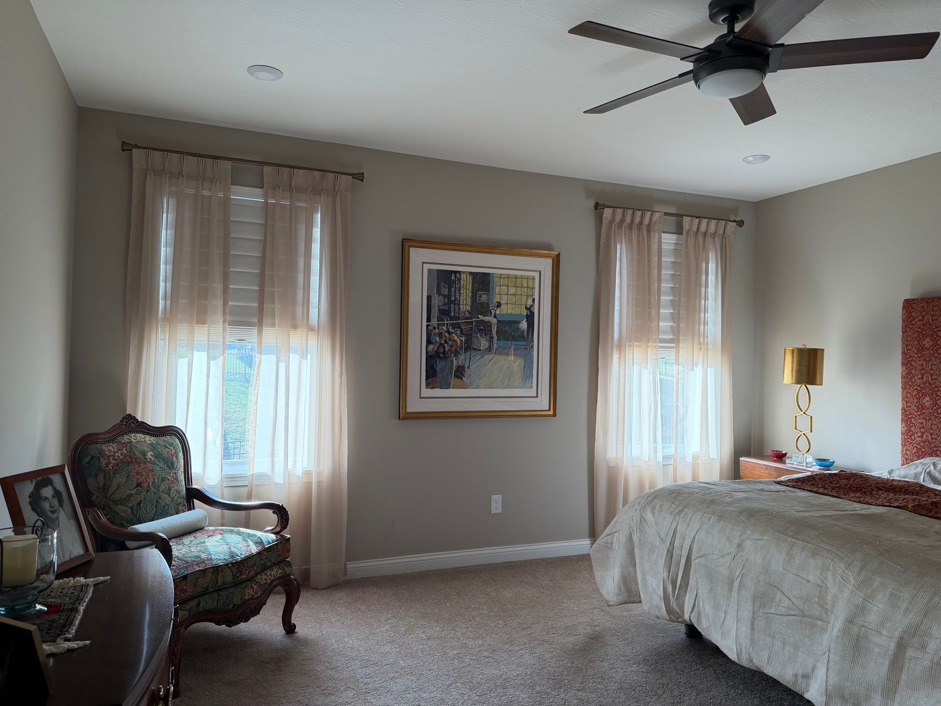 Bedroom with two windows, a bed, armchair, ceiling fan, and framed wall art