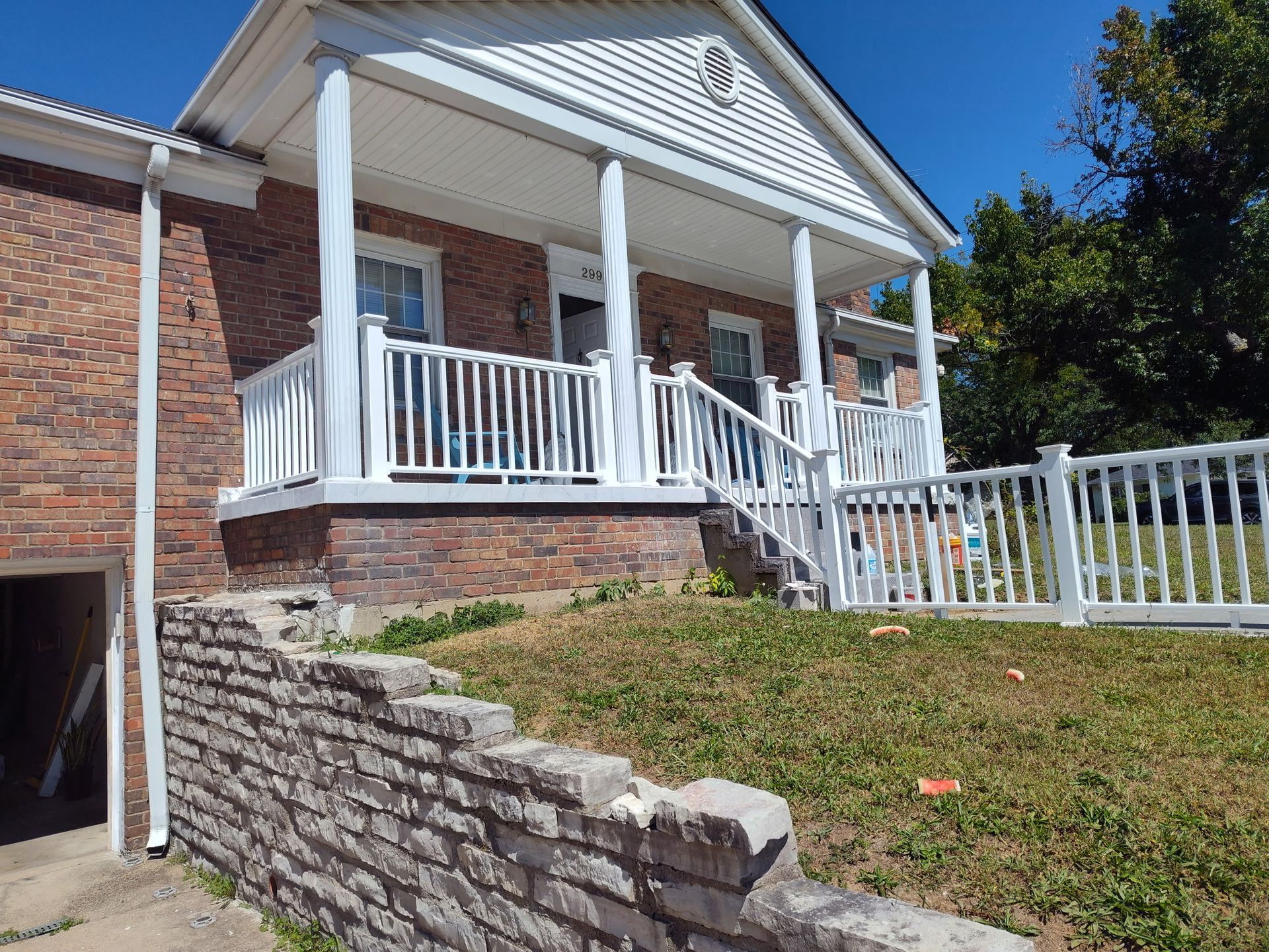 Brick house with white porch and railing, front steps, and a stone retaining wall on a sunny day