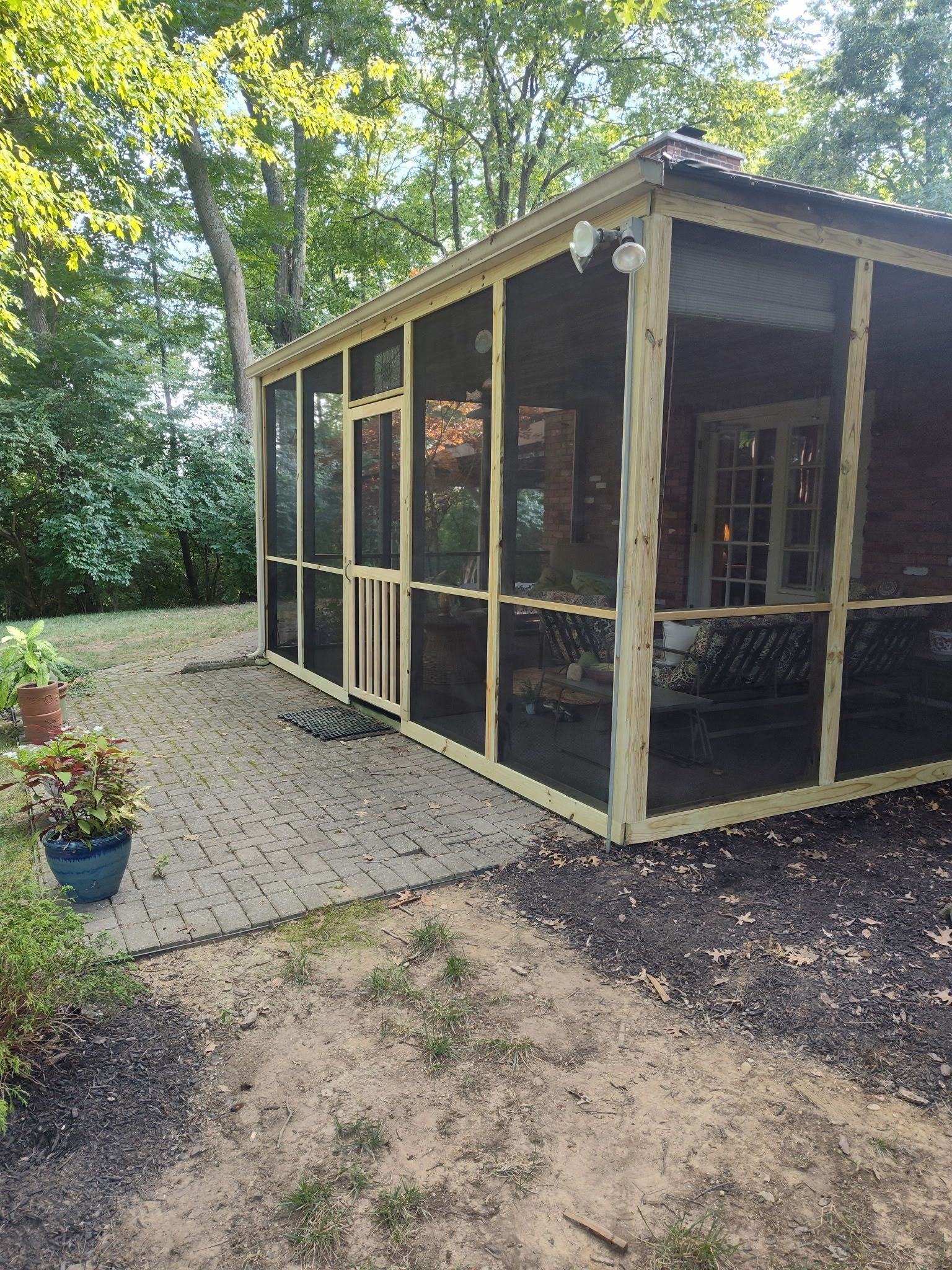 Screened porch with beige trim beside a brick patio, surrounded by trees and greenery