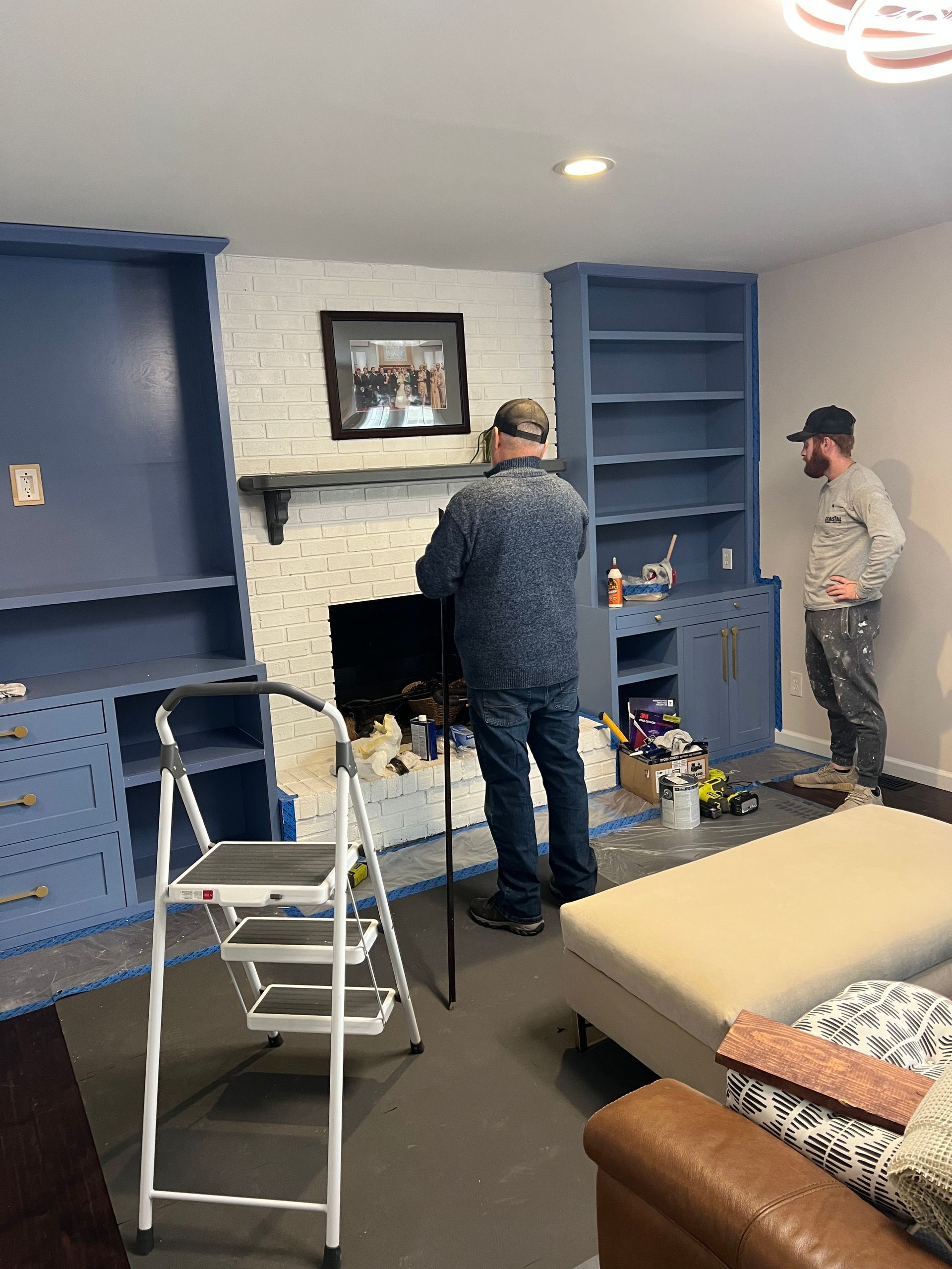 Two people in a blue living room are working on a white brick fireplace with tools and a step stool.