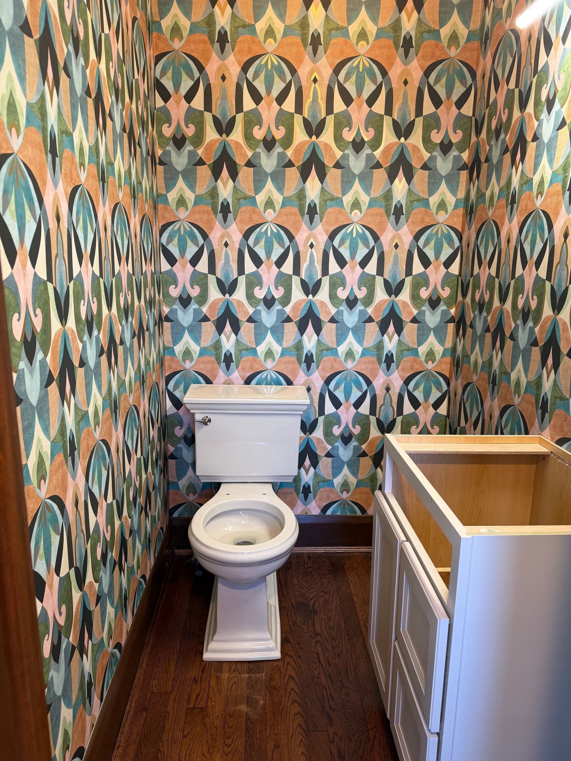 Small bathroom with a toilet, patterned wallpaper, wood floor, and a sink cabinet partially visible.
