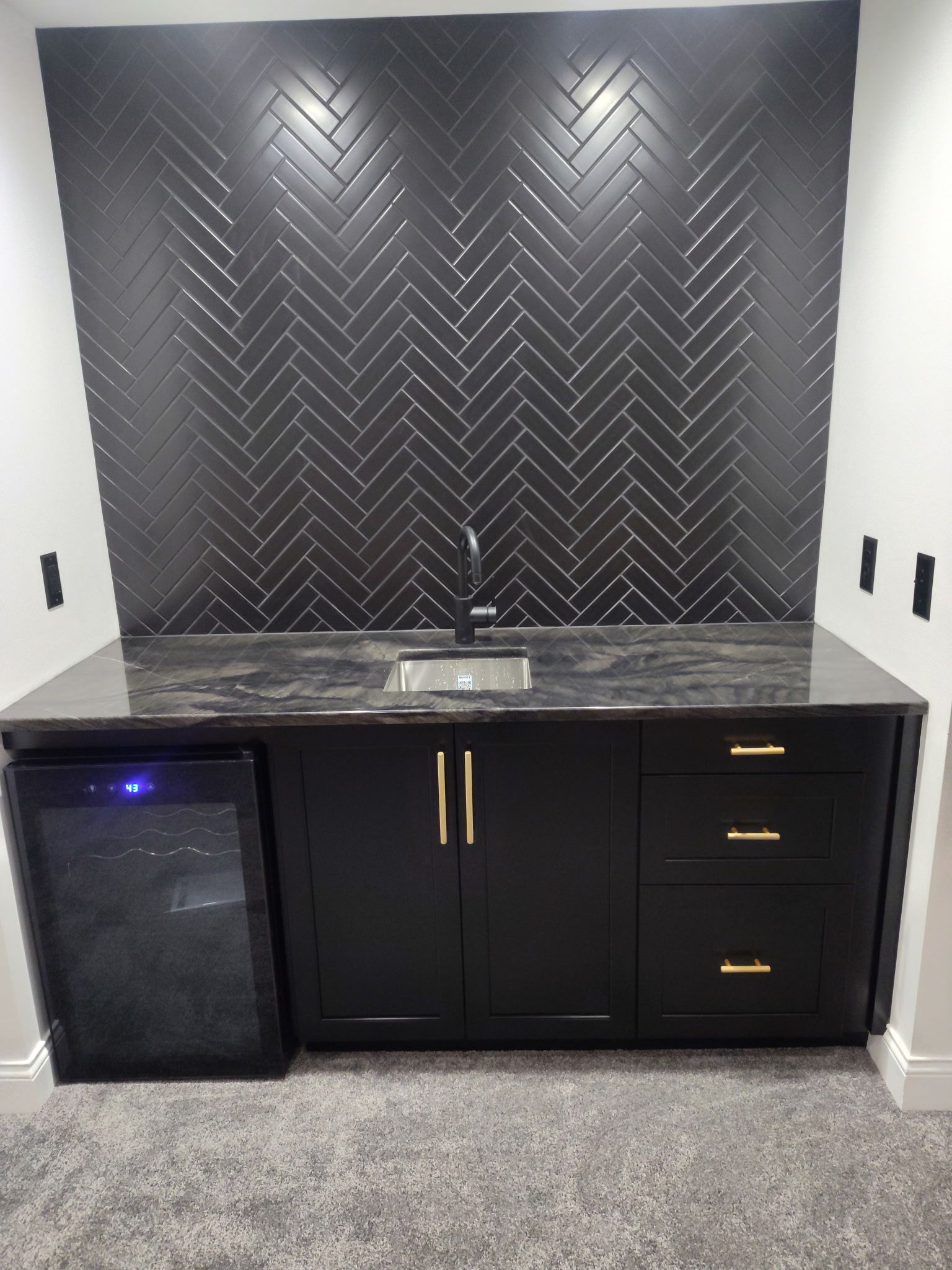 Black wet bar cabinet with mini fridge and drawers beneath a dark herringbone tile backsplash.