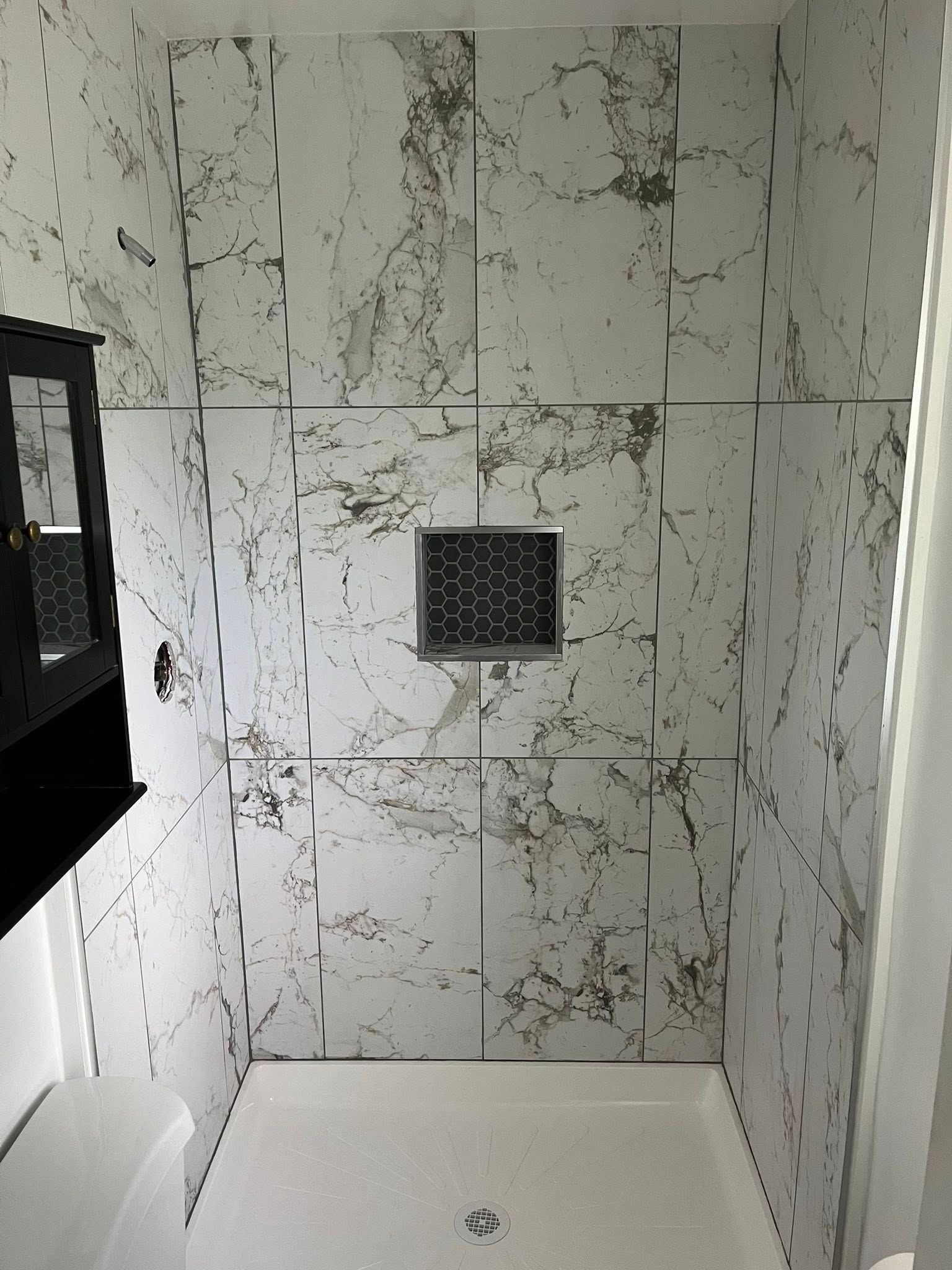 Tiled shower stall with marble-pattern walls, white shower base, and black square vent or niche.
