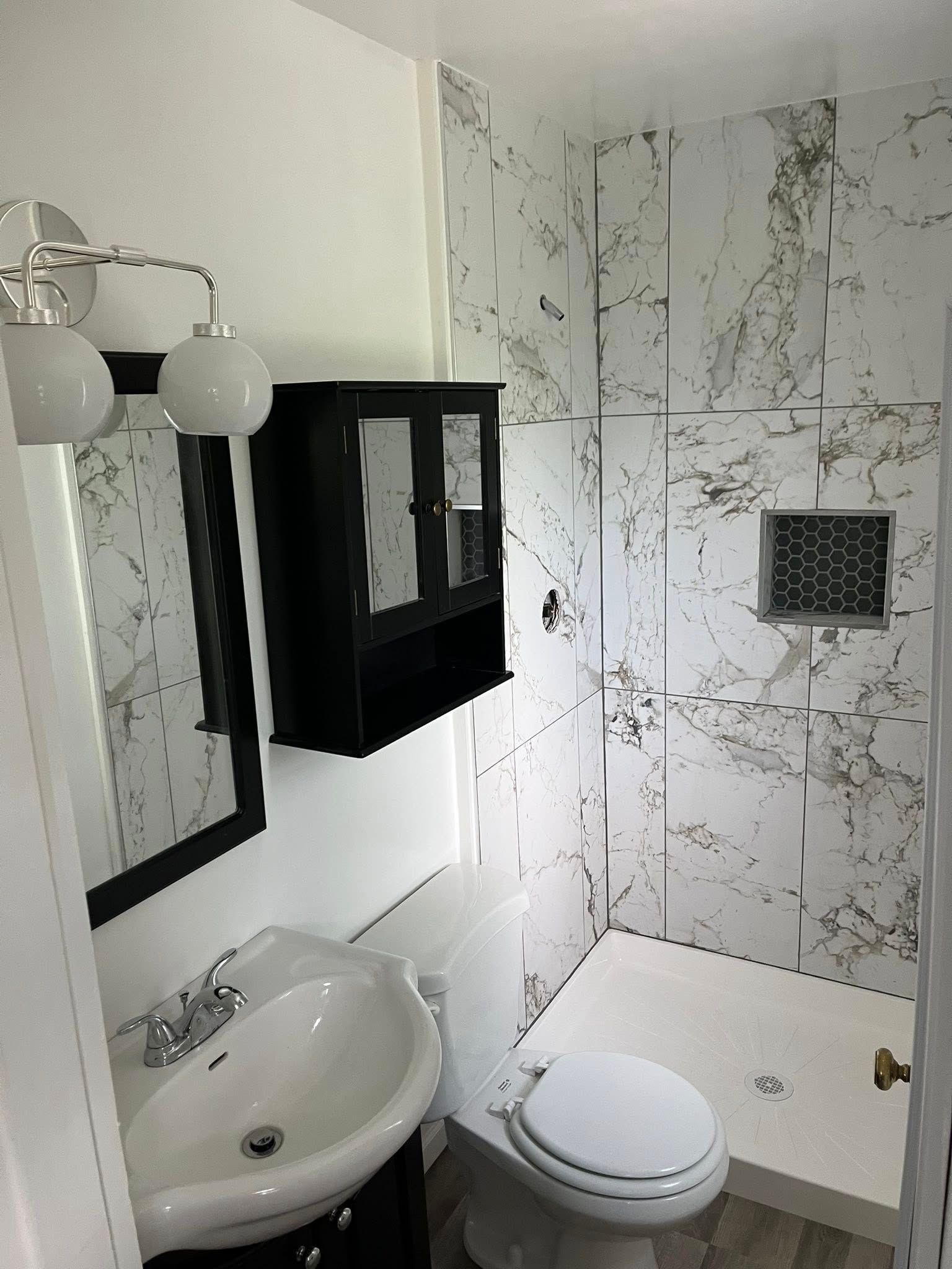 Small tiled bathroom with sink, toilet, shower, mirror, and black wall cabinet.