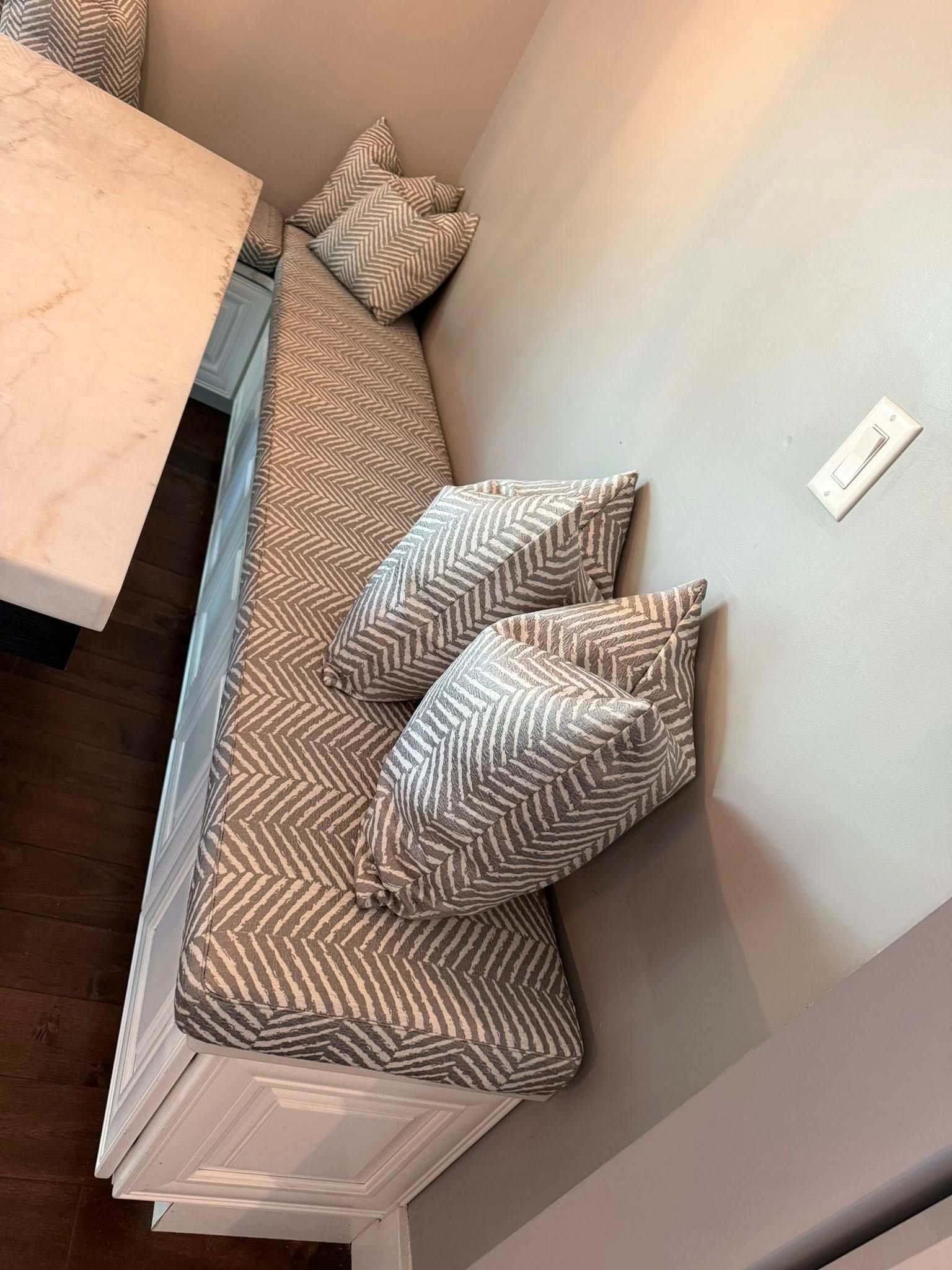 Gray chevron cushioned bench with matching pillows beside a light wall and white side table