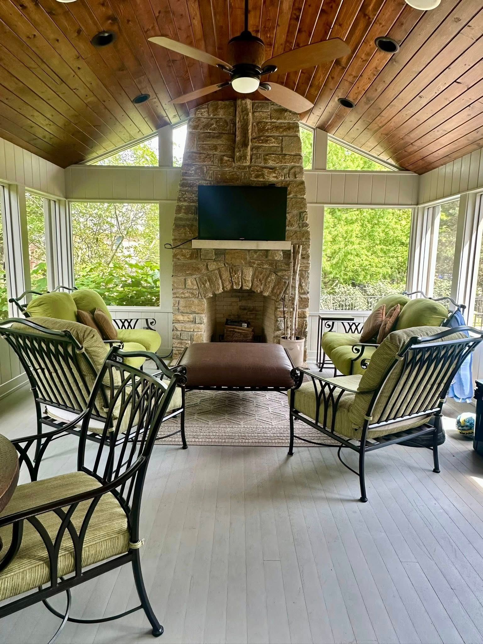 Covered outdoor lounge with wicker chairs, ceiling fan, stone fireplace, and TV.