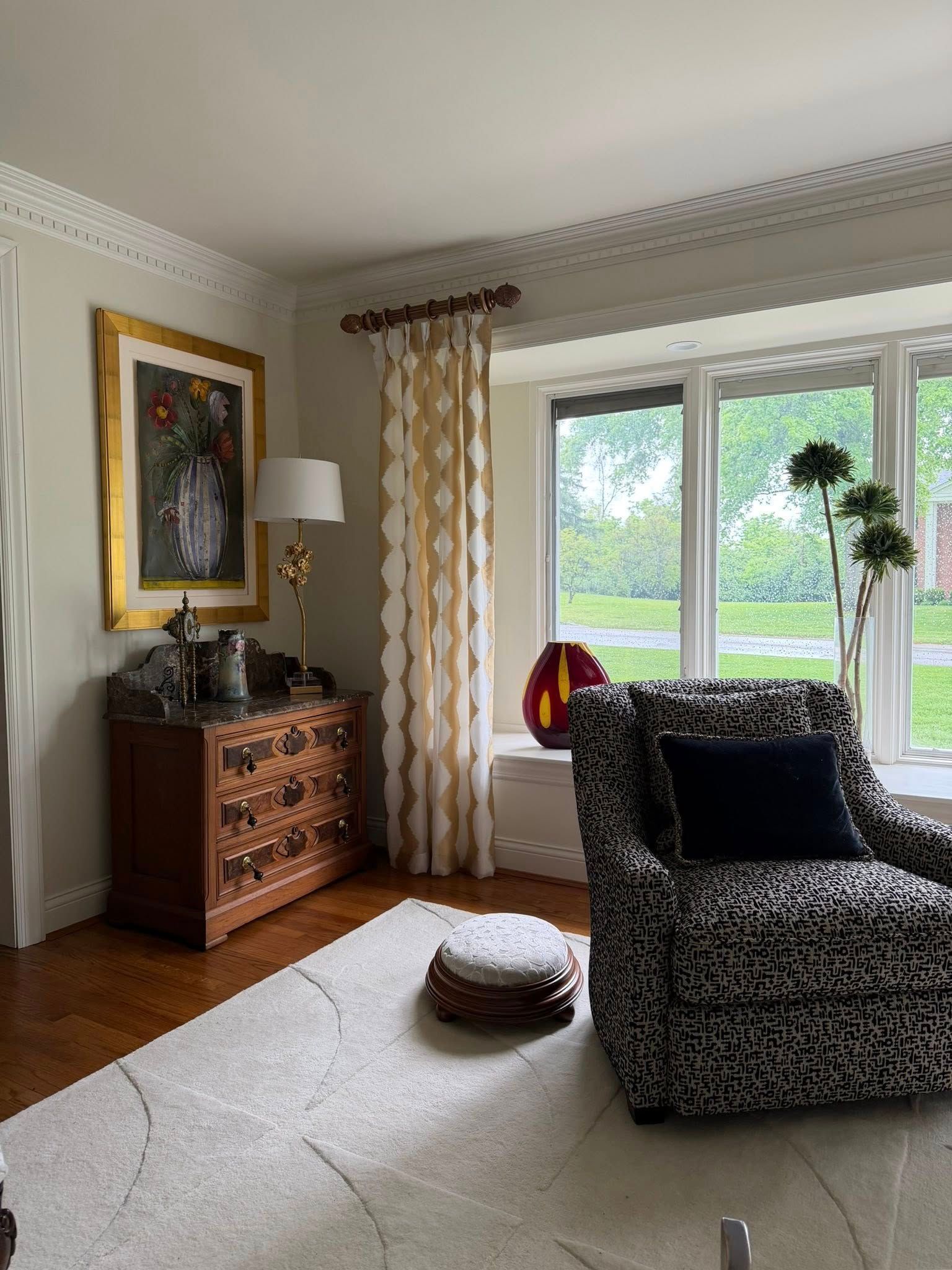 Bright living room with patterned armchair, white rug, wooden chest, curtains, and window view of greenery