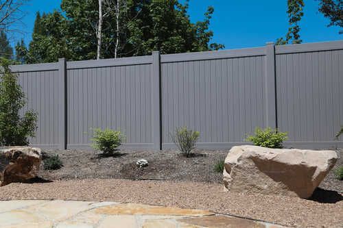 A gray vinyl fence is surrounded by rocks and bushes in a backyard.