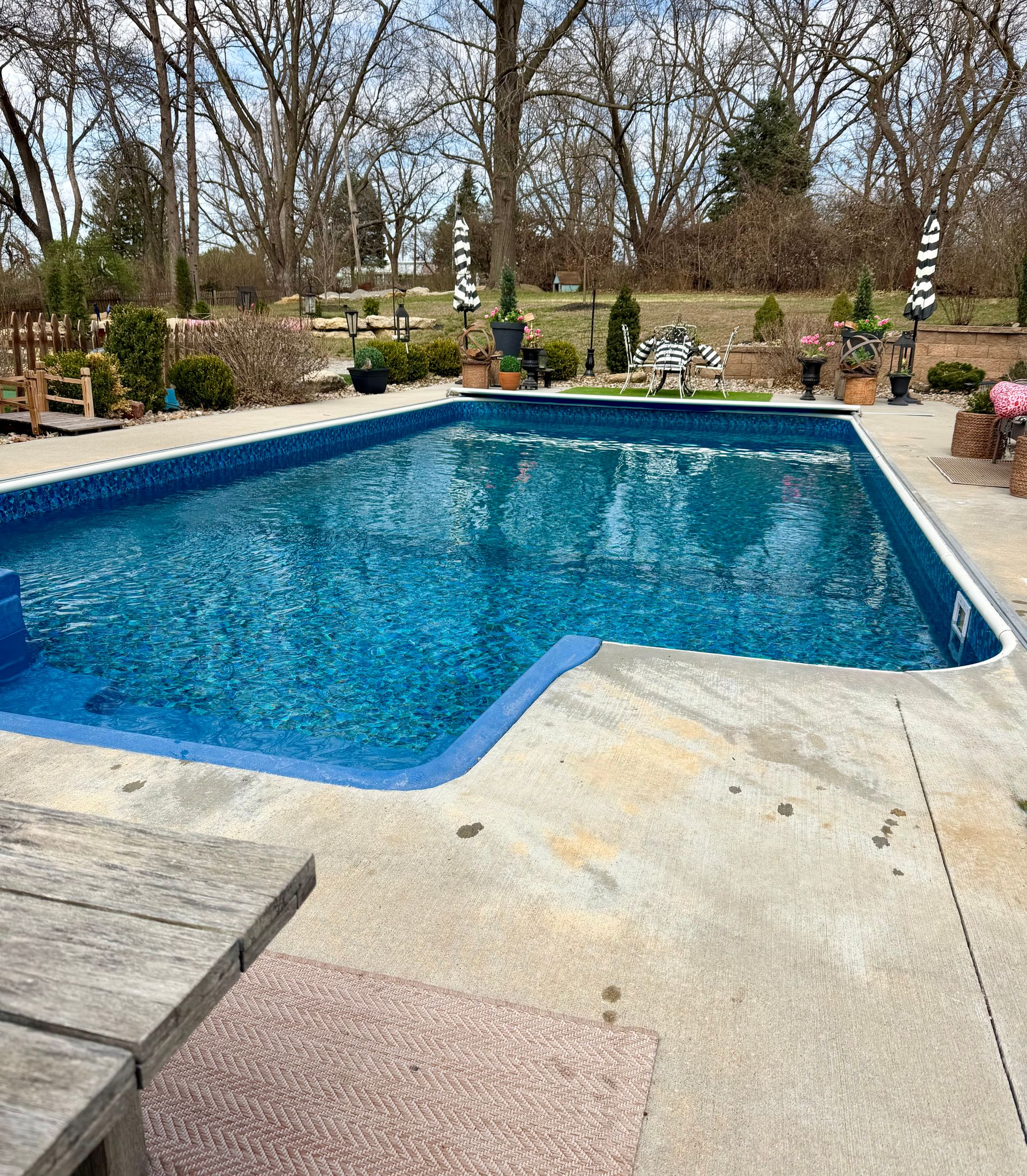 A bright blue rectangular swimming pool surrounded by a concrete deck with a wooden table in the foreground.  