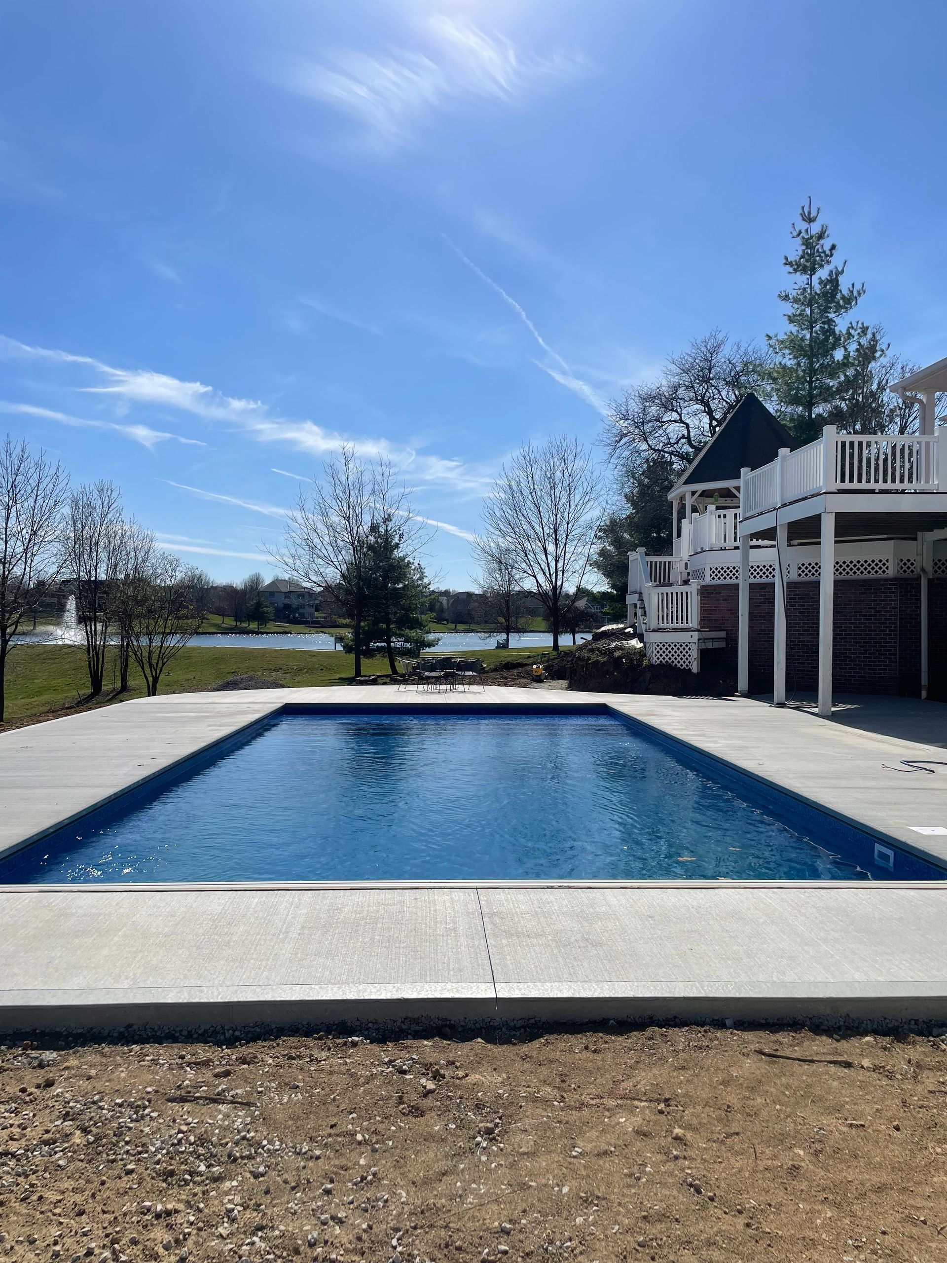 A rectangular swimming pool with blue water and light gray concrete decking, set against a lake and a residential deck. 