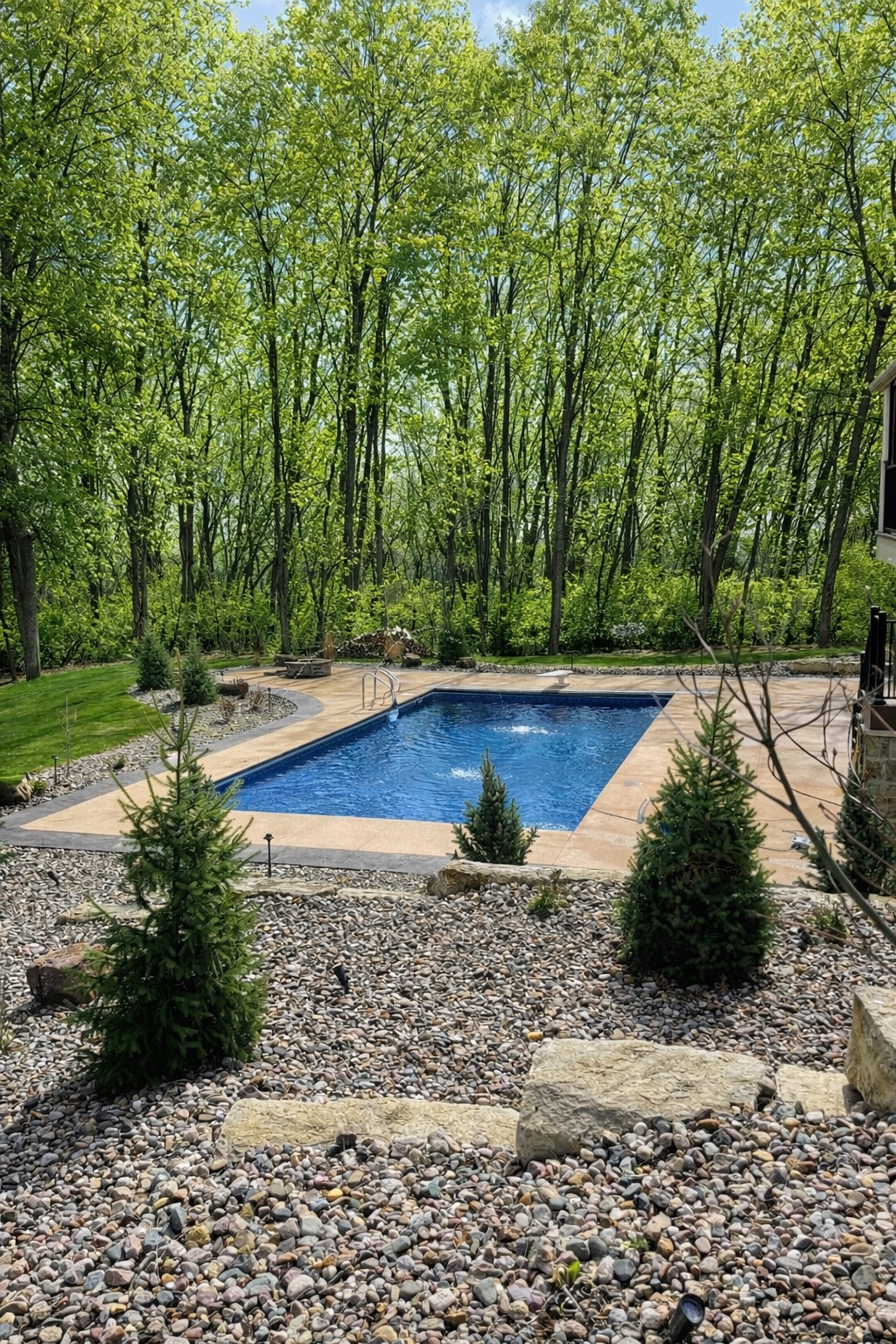 Rectangular outdoor pool with stone border, surrounded by rocks, grass, and trees. Blue water reflects sky.