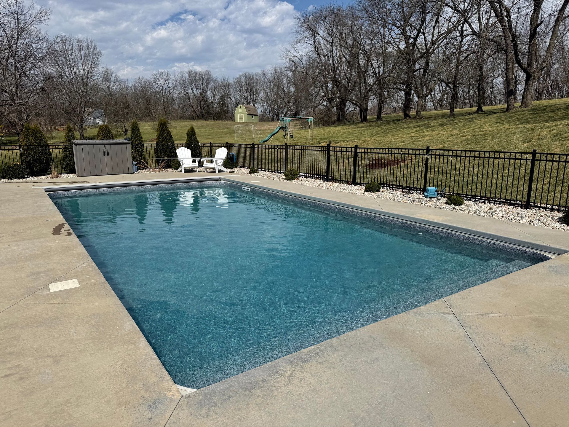 A blue backyard swimming pool surrounded by a concrete deck, with white chairs and a fence in front of a grassy hill. 