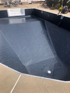 Empty, dark-lined swimming pool with steps, viewed from above. Concrete deck surrounds the pool; construction setting.