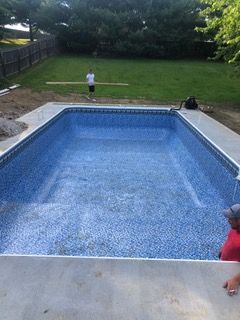 Rectangular in-ground pool, blue mosaic liner, surrounded by concrete. Person stands in yard, another near pool edge.