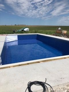 A rectangular blue swimming pool with white steps and concrete surround, under a blue sky and grassy field.