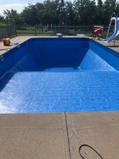 Empty blue swimming pool with concrete surround; a person works in the background.