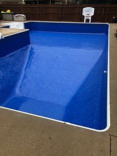 Blue-lined swimming pool, empty, with concrete surround. Wooden fence and white basketball hoop in background.