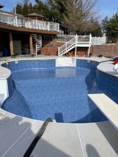 Blue tiled swimming pool with diving board and stairs, surrounded by concrete and a house.