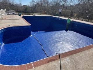 Pool being renovated with workers inside, blue mosaic tiles, concrete deck, and surrounding trees.