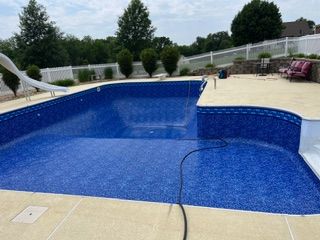 Empty blue swimming pool with concrete deck and slide; surrounded by a white fence and greenery.