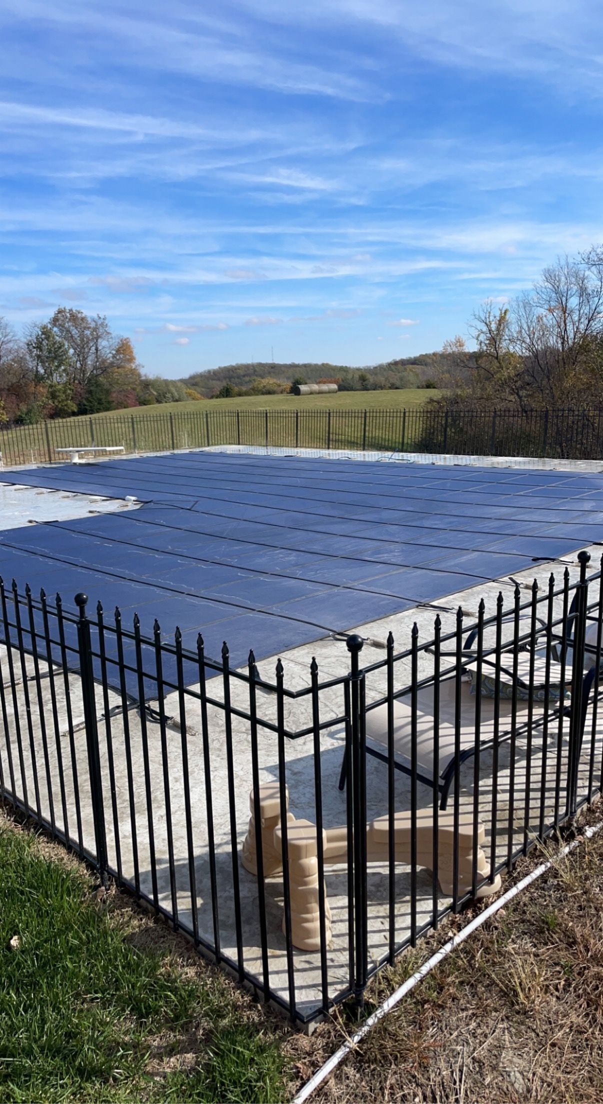 Swimming pool with blue cover, surrounded by black fence, in outdoor setting. Sunny day.