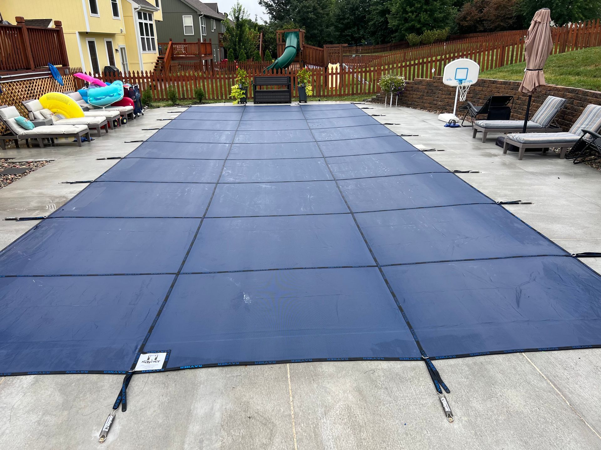 Pool covered with a dark blue safety cover, surrounded by concrete and lounge chairs.