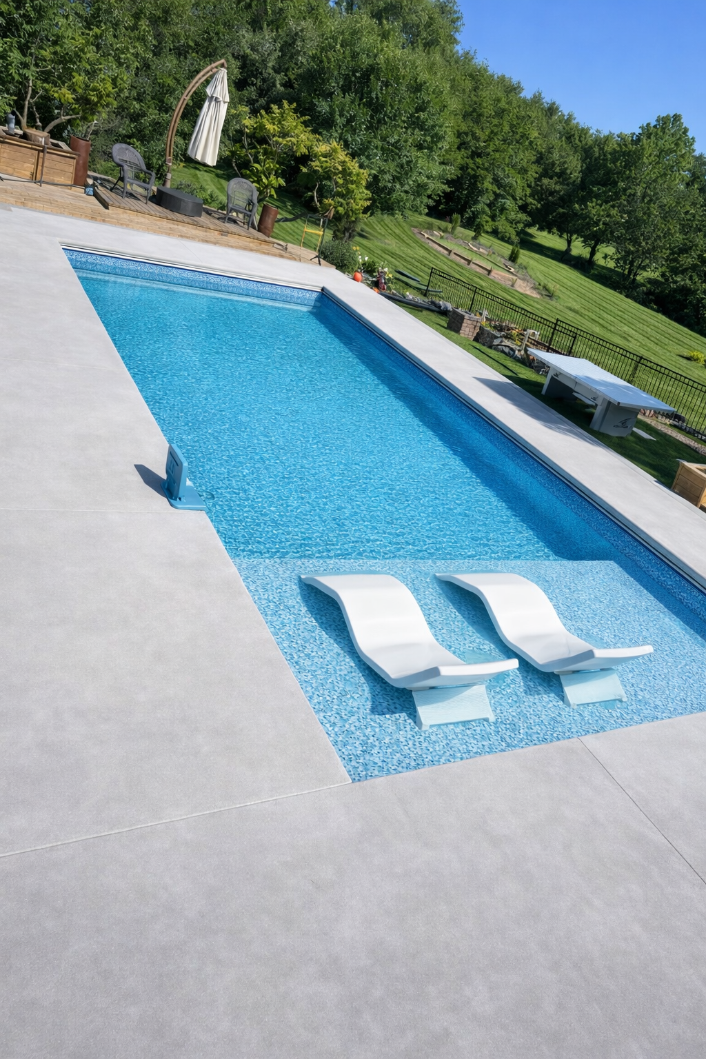 Blue tiled pool with two white lounge chairs submerged. Concrete patio. Green hillside in background.