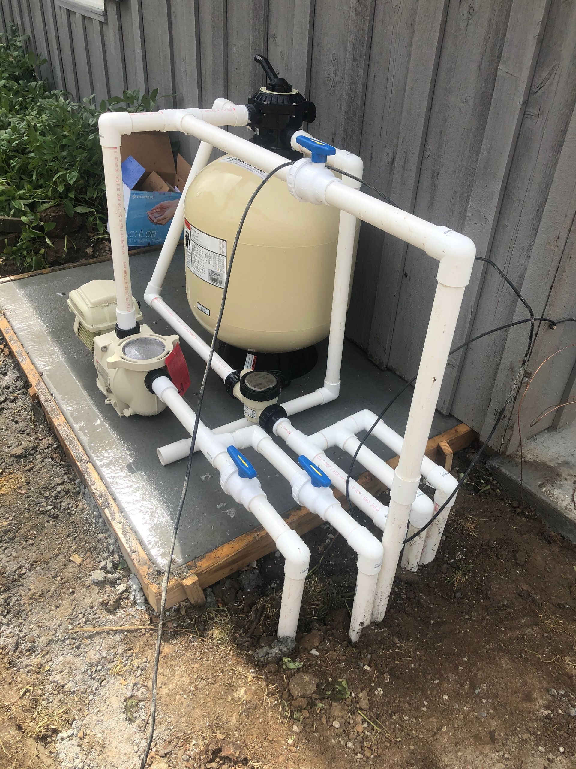 Pool filter system with white pipes, a beige tank, and a pump, outdoors on a concrete pad.