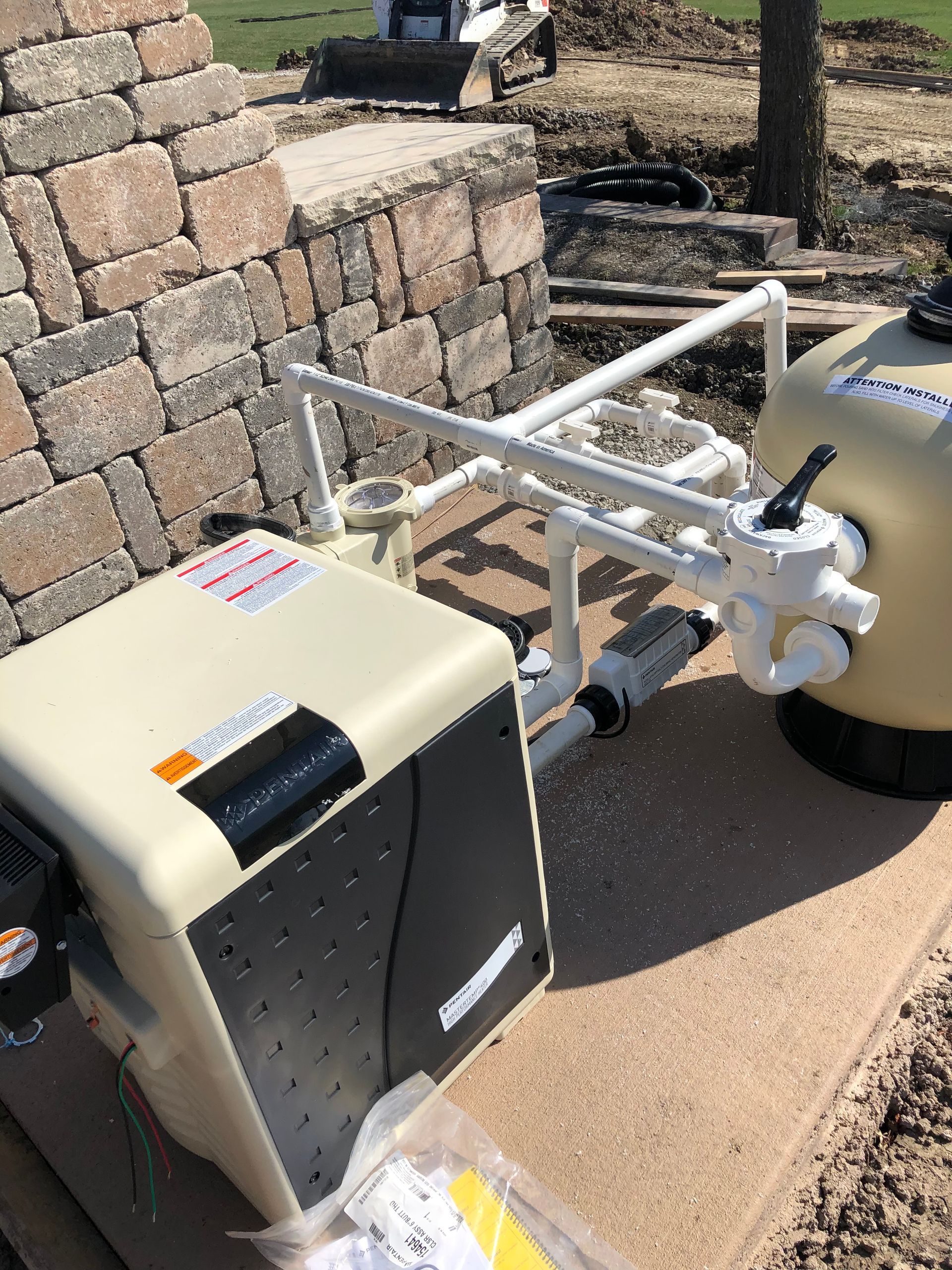Pool equipment: beige heater, filter, pipes on stone patio near a stone wall.