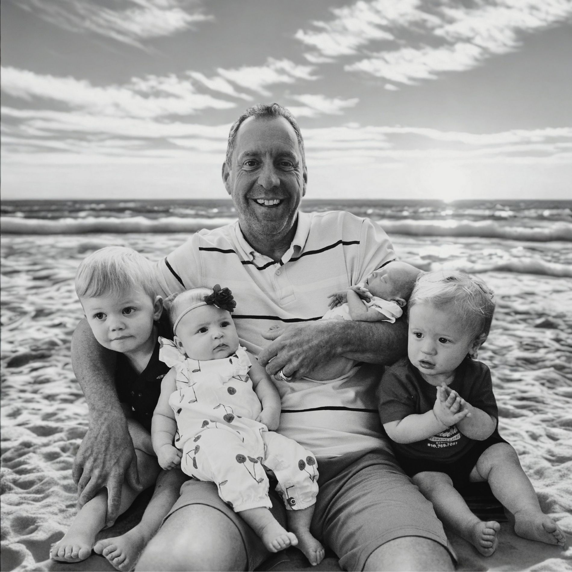 Man on beach with four babies, smiling. Sun and ocean in background. Black and white photo.