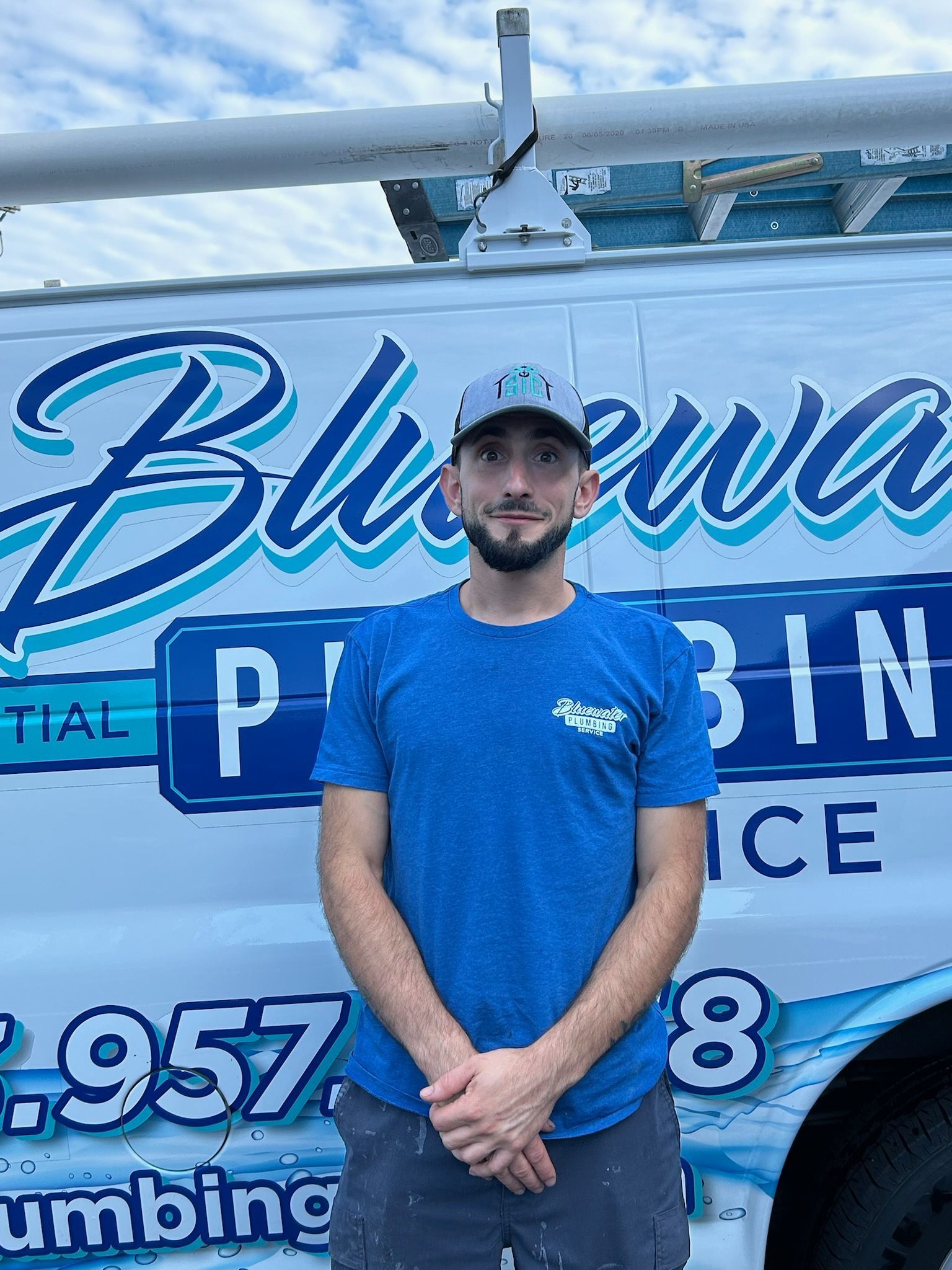 Man in blue shirt and cap stands in front of a blue and white plumbing van.