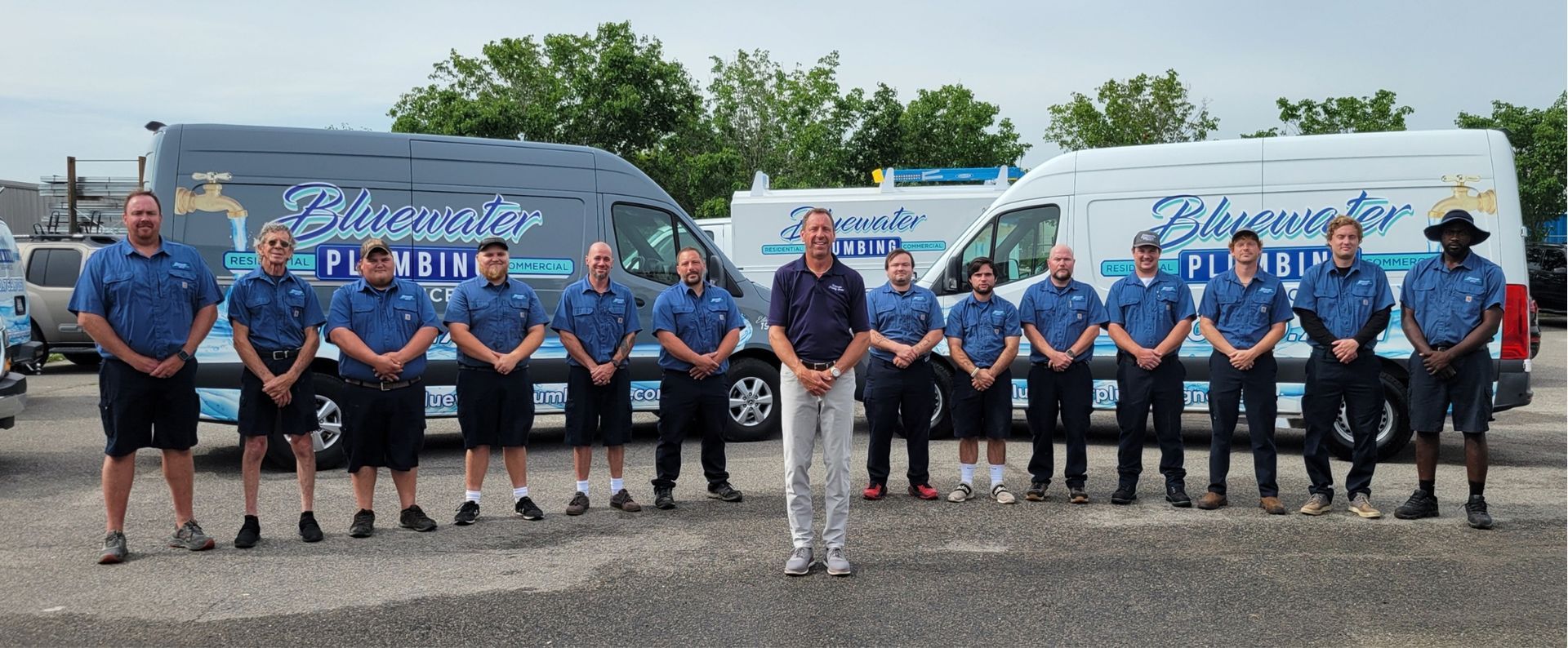 Group of plumbers in blue shirts stand in front of vans with company logo.