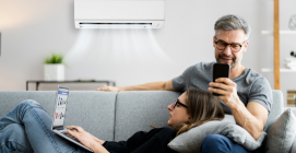 Couple relaxing on couch under air conditioner; one using a laptop, the other with a phone.