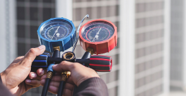 Hands holding a blue and red manifold gauge set; background shows an outdoor air conditioning unit.