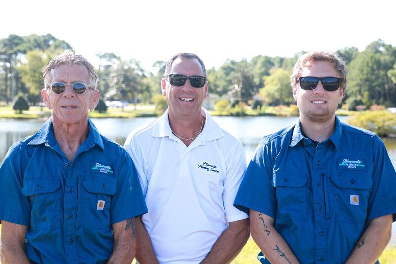 Three men in front of a lake; two in blue work shirts and one in white polo; all wearing sunglasses and smiling.