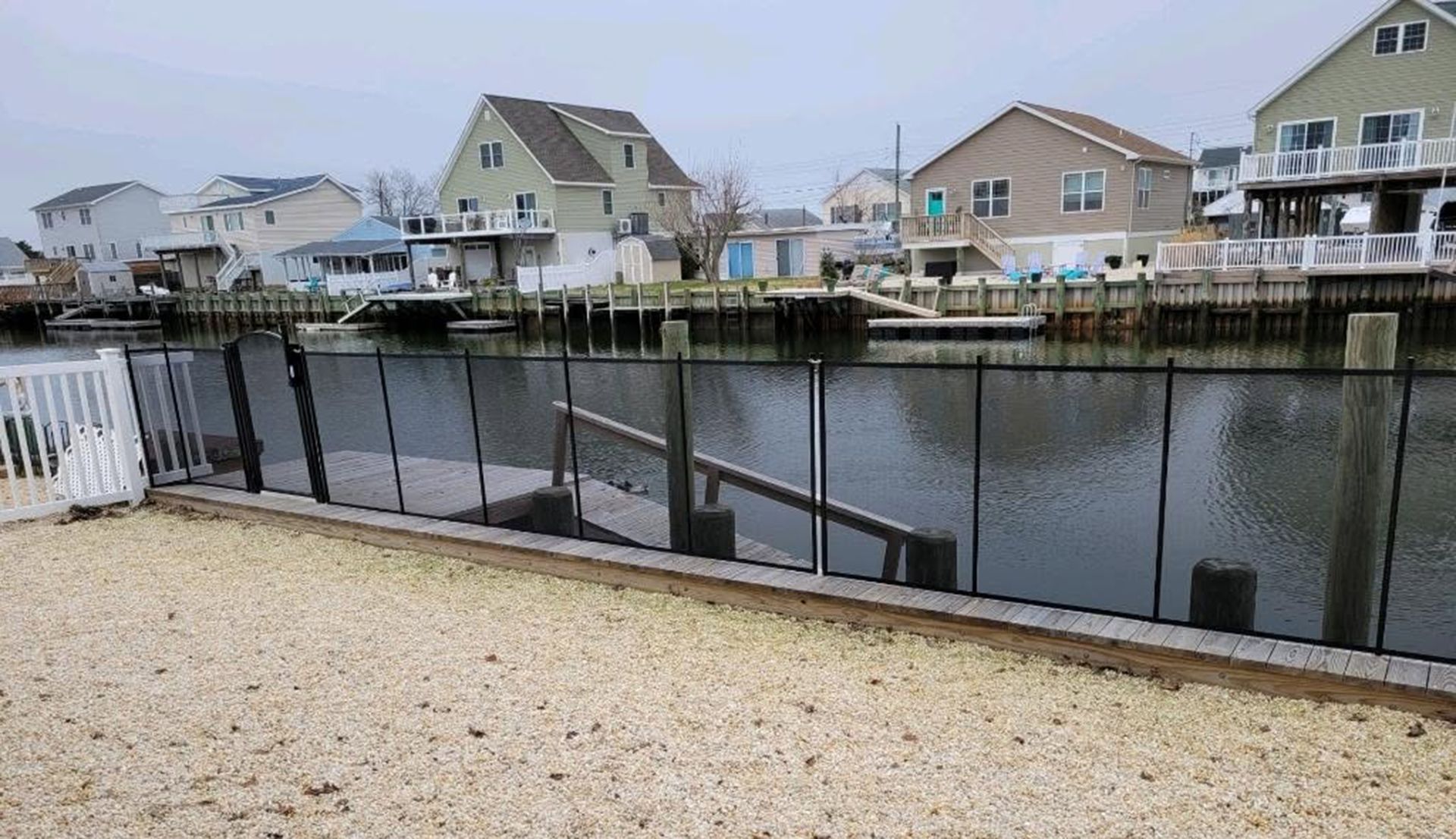 Coastal neighborhood with houses on a canal behind a fenced gravel yard and docked boat slips.